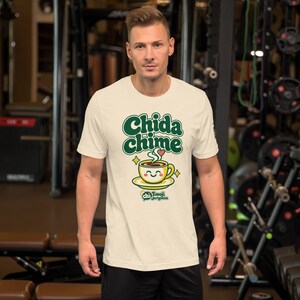 Man in a gym wearing the Heather Natural t-shirt with the Chida chime design: green bubble letters, a smiling coffee cup on a saucer and gold sparkles.