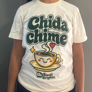 Real photo of the Heather Natural Chida chime t-shirt worn, showing the green retro letters, smiling coffee cup with a coral heart, and the Emoji Jerigonza logo.