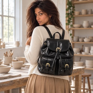 May include: A black leather backpack featuring a top flap with a gold-tone buckle closure, two front pockets with matching gold-tone buckles, a drawstring main compartment, and adjustable shoulder straps, worn over a cream-colored sweater.