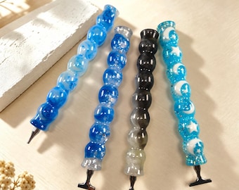 May include: Four resin diamond painting pens with textured, spherical grips. One is light blue with swirls, one is deep blue with glitter, one is translucent black, and one is bright blue with white moon and star shapes embedded inside.