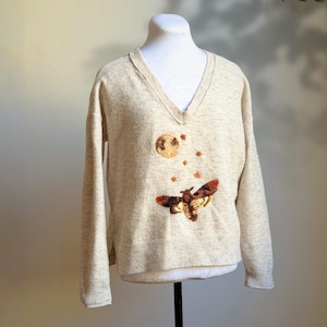 May include: Beige V-neck sweater with an embroidered design featuring a full moon, stars, and a moth. The moth has brown and orange wings. The sweater is displayed on a mannequin.
