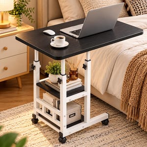 May include: A mobile side table with a black rectangular top and a white metal frame. It features adjustable height knobs, a lower storage shelf, and four black caster wheels. The table holds a laptop, a white coffee cup, and a computer mouse.