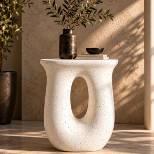 May include: A white, speckled, hourglass-shaped side table with a large, central oval cutout. The table features a smooth, curved silhouette and a wide, flat top surface. It is constructed from a solid material with a matte, textured finish.