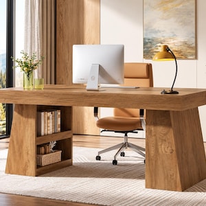 May include: A wooden desk with a thick rectangular top and two wide, angled support legs. The left leg features two open shelves containing books and a woven basket. A white computer monitor, a yellow desk lamp, and a tan office chair.