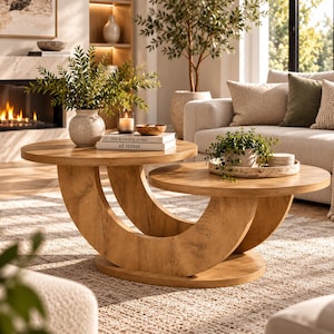 May include: A round wooden coffee table featuring two circular tabletops supported by a curved, crescent-shaped base. The table has a light brown wood grain finish and is topped with a ceramic vase, a candle, and a book titled The Kinfolk Home.