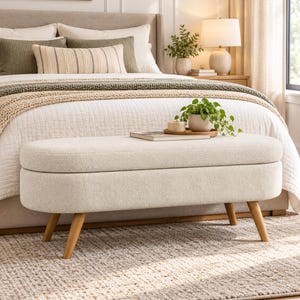 May include: An oval-shaped storage bench upholstered in cream-colored textured fabric, featuring a hinged lid and four tapered light wood legs. A small potted green plant and a thin book rest on the flat top surface of the bench.