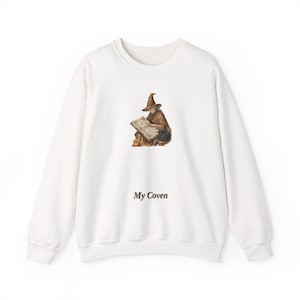 May include: A white long-sleeve sweatshirt featuring a central graphic of a figure in a pointed hat reading an open book with a black cat nearby. Below the illustration, the text "My Coven" is printed in a dark, simple serif font.