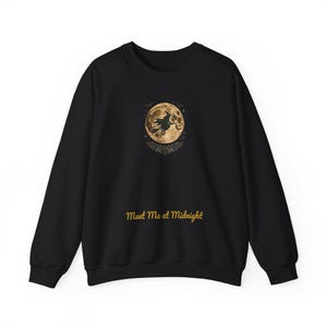 May include: A black long-sleeve sweatshirt featuring a central graphic of a full moon with a silhouette of a witch flying on a broomstick, surrounded by small bats and foliage. Below the graphic, the text reads Meet Me at Midnight in gold.