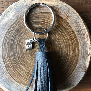 May include: A black leather tassel keyring with a silver key ring and a small charm with the letters ABC. The keyring is displayed on a wooden slice with visible growth rings.