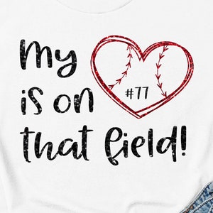 May include: White t-shirt with black text that reads "My heart is on that field!" and a red glitter heart with a baseball design and the number "#77" inside. The text "Iron on transfer only!" is at the bottom of the image.