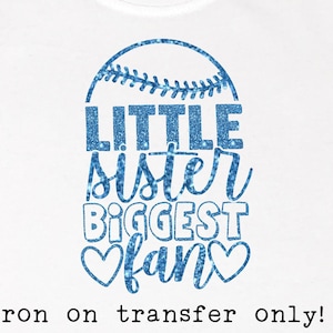 Puede incluir: Camiseta blanca con un diseño de béisbol azul brillante. El diseño incluye las palabras "LITTLE SISTER BIGGEST FAN" y dos corazones. La frase "iron on transfer only!" está impresa en la parte inferior.