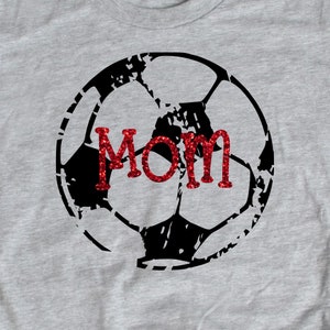 Może przedstawiać: Szara koszulka z nadrukiem czarno-białej piłki nożnej. Słowo "MAMA" jest napisane czerwonym brokatem na piłce nożnej.