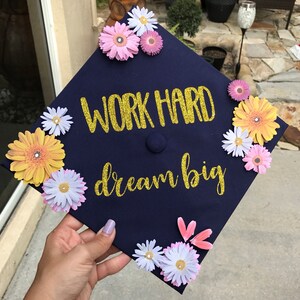 K&ouml;nnte beinhalten: Eine marineblaue Abschluss-Kappe, verziert mit Papierblumen in Rosa-, Gelb- und Wei&szlig;t&ouml;nen. Die Kappe tr&auml;gt goldene Glitzerbuchstaben mit der Aufschrift "Work Hard Dream Big".