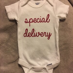 K&ouml;nnte beinhalten: Wei&szlig;er Baby-Strampler mit dem Schriftzug "special delivery" in roten Glitzerbuchstaben.