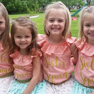 K&ouml;nnte beinhalten: Vier junge M&auml;dchen tragen pfirsichfarbene Shirts mit goldenen Glitzerbuchstaben, auf denen "Sister Squad" steht. Die M&auml;dchen tragen verschiedene farbige R&ouml;cke, eines tr&auml;gt einen wei&szlig;en Rock, eines tr&auml;gt einen hellblauen Rock und zwei tragen einen t&uuml;rkisfarbenen Rock. Die M&auml;dchen l&auml;cheln und schauen in die Kamera.