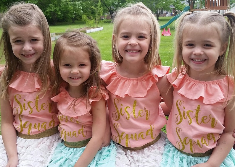 K&ouml;nnte beinhalten: Vier junge M&auml;dchen tragen pfirsichfarbene Shirts mit goldenen Glitzerbuchstaben, auf denen "Sister Squad" steht. Die M&auml;dchen tragen verschiedene farbige R&ouml;cke, eines tr&auml;gt einen wei&szlig;en Rock, eines tr&auml;gt einen hellblauen Rock und zwei tragen einen t&uuml;rkisfarbenen Rock. Die M&auml;dchen l&auml;cheln und schauen in die Kamera.