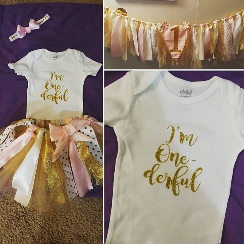 K&ouml;nnte beinhalten: Ein wei&szlig;er Strampler mit goldenen Glitzerbuchstaben, auf dem "I'm One-derful" steht. Der Strampler wird mit einem T&uuml;ll-Tutu mit rosa, goldenen und wei&szlig;en B&auml;ndern kombiniert. Ein rosa Stirnband mit Schleife ist ebenfalls enthalten.