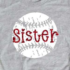 Puede incluir: Una camiseta gris con un gráfico de béisbol blanco y la palabra "Hermana" en letras de purpurina roja.