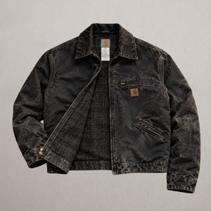 May include: A black, distressed-finish jacket with a fold-down collar, a full-length front zip, and a plaid-lined interior. It features a zipped chest pocket, two lower zipped pockets, and a square Carhartt brand patch on the chest.