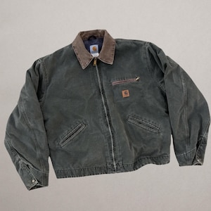Puede incluir: Una chaqueta de lona verde bosque oscuro con cuello de pana marrón, cremallera frontal completa y mangas largas con puños abotonados. La prenda incluye un bolsillo en el pecho con cremallera, dos bolsillos inferiores y un parche de la marca Carhartt.