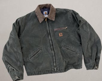 Può includere: Una giacca in tela verde bosco scuro con colletto in velluto a coste marrone, chiusura frontale con zip integrale e maniche lunghe con polsini abbottonati. Il capo presenta una tasca sul petto con zip, due tasche a filetto e un logo Carhartt.