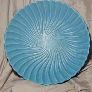 May include: A round, light blue ceramic plate featuring a textured surface with a radiating spiral pattern that extends from the center to the scalloped, wavy edges. The finish has a matte, slightly speckled appearance across the entire surface.