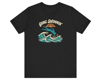 May include: A black short-sleeve t-shirt featuring a graphic print of a person diving into ocean waves. Above the swimmer is a stylized orange sun and the text GONE SWIMMIN' in a cream-colored retro font. The waves are depicted in shades of teal.