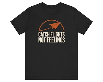 May include: A black short-sleeve t-shirt featuring a graphic of an orange paper airplane flying over a curved line. Below the graphic, the text reads CATCH FLIGHTS NOT FEELINGS in a bold, off-white, sans-serif font centered on the chest area.