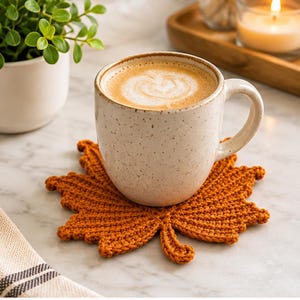 Puede incluir: Una taza de cerámica color crema con motas llena de café descansa sobre un posavasos de ganchillo de color naranja vibrante con forma de hoja de arce. El posavasos presenta un tejido de hilo intrincado y un tallo distintivo en la base.
