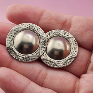 May include: A pair of circular silver-toned earrings featuring a smooth, domed center surrounded by a decorative border with intricate engraved scrollwork and small floral motifs. The earrings have a polished metallic finish and a round shape.
