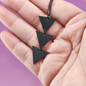 May include: A necklace featuring three matte black metal triangles arranged in a vertical line. The triangles are connected by small black metal jump rings and suspended from a thin black metal chain, creating a geometric pendant design.