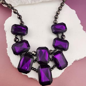 May include: A statement necklace featuring seven large, rectangular, deep purple faceted stones arranged in a tiered cluster. The stones are connected by dark metallic, gunmetal-colored beads that form the chain and link the segments.