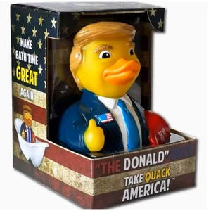 May include: A yellow rubber duck shaped like a person with blonde hair, wearing a blue suit and white shirt with a blue tie. The packaging features the text "MAKE BATH TIME GREAT AGAIN" and "THE DONALD TAKE QUACK AMERICA!" with American flag motifs.