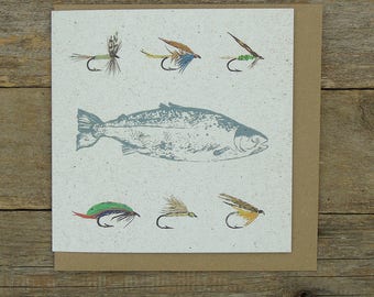 May include: A square greeting card featuring a fish illustration and six fishing flies in different colours. The card is on a brown envelope, set against a wooden background. The fish is a light blue colour.