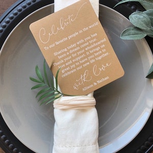 May include: A brown card with white text that reads "Celebrate" and "To our favorite people in the world! Sharing today with you has been a dream come true. Thank you for your wholehearted love and support. It brings us great joy to have you here to celebrate our new life together. With love, Allison & Nathan." The card is placed on a white napkin with a green leaf and a brown ribbon. The napkin is on a gray plate with a black rim.