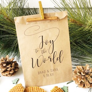 May include: A brown paper bag with a cinnamon stick and a gold clip. The bag has the text "Joy to the World" with the names "Brad & Sara" and the date "5.18.XX" printed on it.