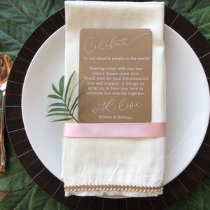 May include: A brown card with white text that reads "Celebrate" and "To our favorite people in the world! Sharing today with you has been a dream come true. Thank you for your wholehearted love and support. It brings us great joy to have you here to celebrate our new life together. With love, Allison & Nathan". The card is tied with a pink ribbon and placed on a white linen napkin.