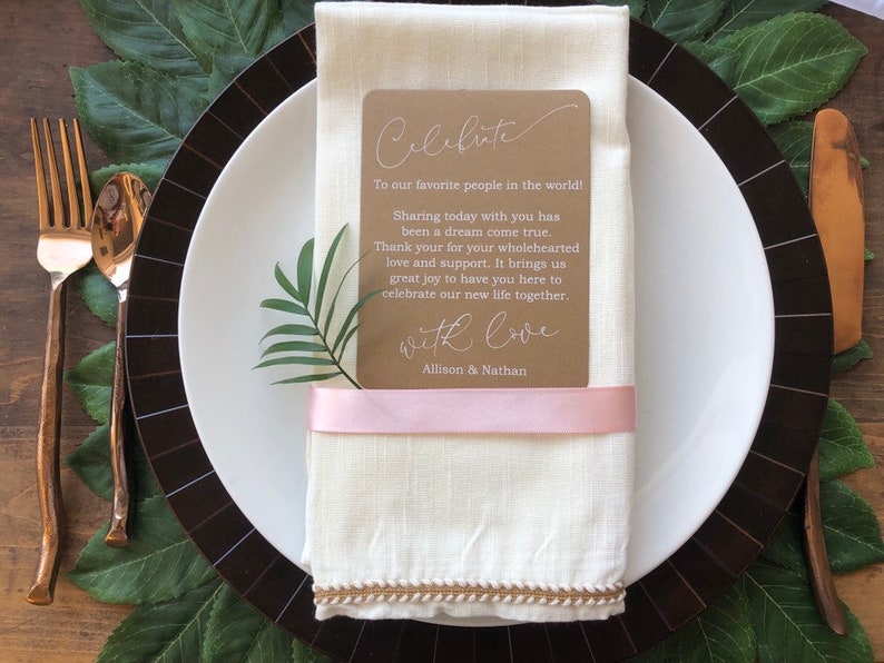 May include: A brown card with white text that reads "Celebrate" and "To our favorite people in the world! Sharing today with you has been a dream come true. Thank you for your wholehearted love and support. It brings us great joy to have you here to celebrate our new life together. With love, Allison & Nathan". The card is tied with a pink ribbon and placed on a white linen napkin.