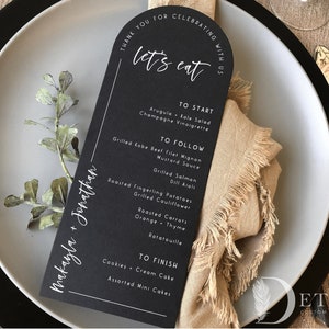 May include: Black and white wedding menu card with the text "Thank you for celebrating with us" and "Let's eat". The menu includes a list of dishes for a wedding reception, including arugula kale salad, grilled Kobe beef filet mignon, grilled salmon, roasted fingerling potatoes, and assorted mini cakes. The card is on a white plate with a brown napkin.