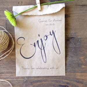 May include: A brown paper bag with a green flower and a white clothes peg attached to the top. The bag has the text "Garret & Amber 04.23.16" and "Enjoy" written on it. The bag also has the text "Thanks for celebrating with us!" written on it.