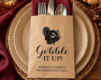 May include: A brown paper cutlery pouch containing a silver fork, knife, and spoon. The pouch features a black graphic of a turkey above the text "Gobble IT UP!" and "JOHNSON FAMILY THANKSGIVING 20xx" printed in black ink on the front surface.