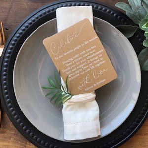 May include: A brown card with white text that reads "Celebrate" and a message about sharing a special day with loved ones. The card is tied to a white napkin with a brown twine bow. The napkin is on a gray plate with a black rim.