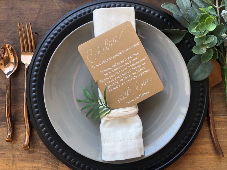 May include: A brown card with white text that reads "Celebrate" and a message about sharing a special day with loved ones. The card is tied to a white napkin with a brown twine bow. The napkin is on a gray plate with a black rim.