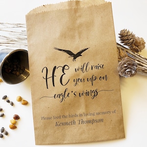 Può includere: Un sacchetto di carta marrone con una silhouette nera di un'aquila e il testo "HE will raise you up on eagle's wings". Sotto il testo si legge "Please feed the birds in loving memory of Kenneth Thompson".