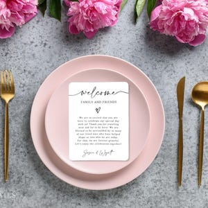 K&ouml;nnte beinhalten: Eine Hochzeitstafel mit rosa Tellern, goldenem Besteck und einer Willkommenskarte. Auf der Karte steht "Willkommen Familie und Freunde" mit einem Herzsymbol und einer Nachricht. Rosa Pfingstrosen sind um die Teller angeordnet.