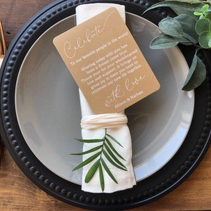 May include: A black and gray charger plate with a white napkin tied with a brown card. The card has the text "Celebrate" and a message thanking guests for their support. The card is tied with a white ribbon and a green leaf is tucked into the napkin.