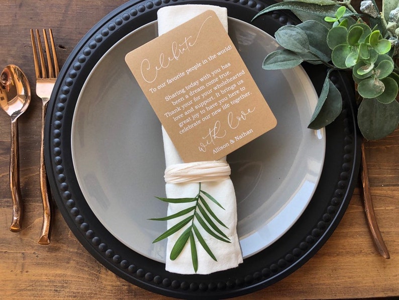 May include: A black and gray charger plate with a white napkin tied with a brown card. The card has the text "Celebrate" and a message thanking guests for their support. The card is tied with a white ribbon and a green leaf is tucked into the napkin.