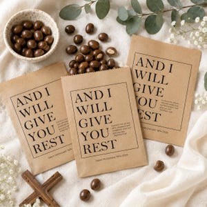 Può includere: Tre sacchetti rettangolari in carta marrone con il testo stampato in nero AND I WILL GIVE YOU REST, una citazione biblica più piccola e il nome Jason McDonald 1974-2025. Una ciotola in ceramica e noci ricoperte di cioccolato sono vicine.