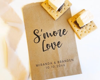 Puede incluir: Una bolsa de papel marrón con las palabras "S'more Love" en escritura negra. La bolsa también incluye los nombres "Miranda & Branden" y la fecha "10.10.20XX". Dos s'mores de galletas Graham están al lado de la bolsa.