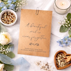 Può includere: Un sacchetto di semi di carta marrone con la scritta "A beautiful soul is never forgotten". Una graffetta argentata fissa la parte superiore. Il sacchetto è circondato da fiori bianchi, fiori blu, una candela e bustine di semi.